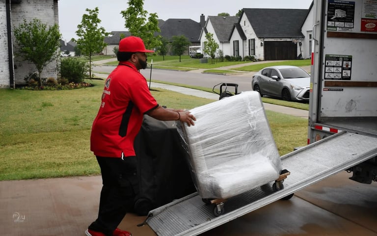 A mover loads a wrapped chair onto a truck ramp in a suburban neighborhood.