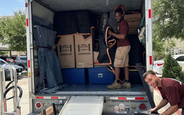 Two movers loading furniture into a moving truck on a sunny day.