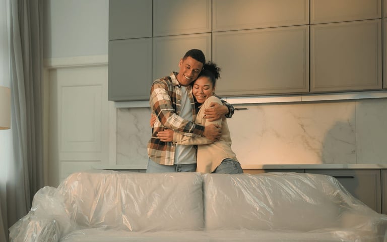 Couple embraces in new home with sofa wrapped in plastic.