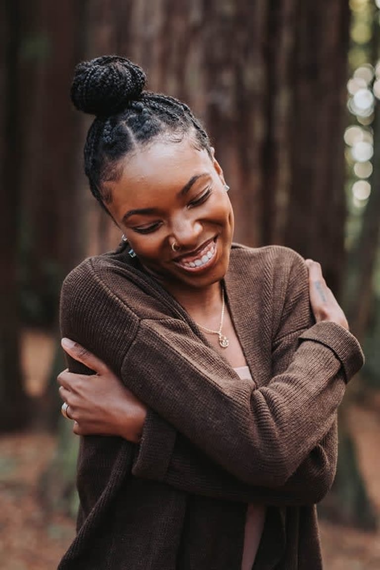A woman in a forest hugging herself with a joyful expression.