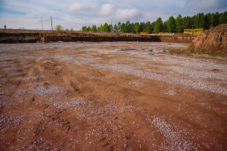 An empty dirt lot prepared for construction with visible tire tracks and distant machinery.