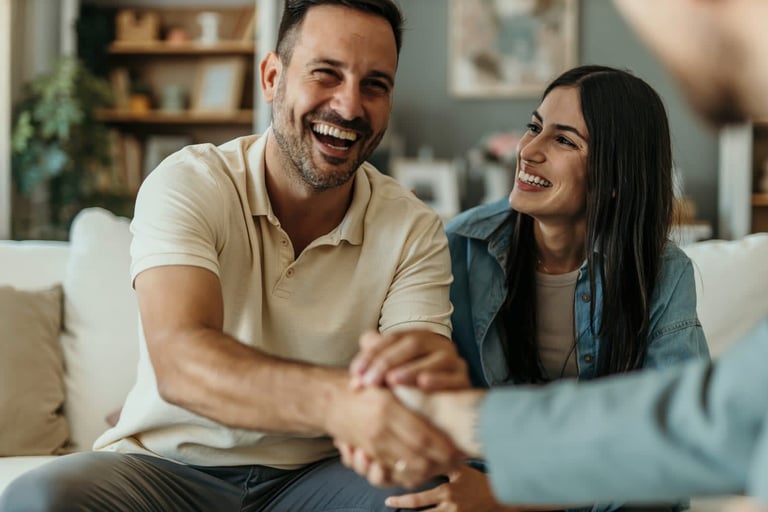 Two people laughing while shaking hands on a couch in a living room.