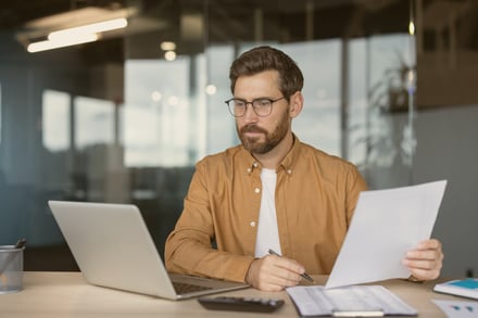 Man reviewing documents at a desk with laptop and calculator.