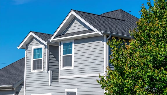 Gray house with gable roof, dark gray shingles, white window frames, gray vinyl siding, and a green leafy tree partially obscuring the view against a bright blue sky. The image shows the upper portion