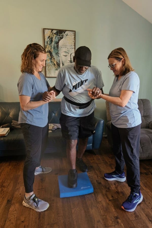 Three people in athletic wear guiding a man on a blue step platform indoors.