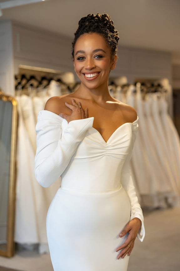 Woman in white off-the-shoulder wedding dress smiling in bridal shop.