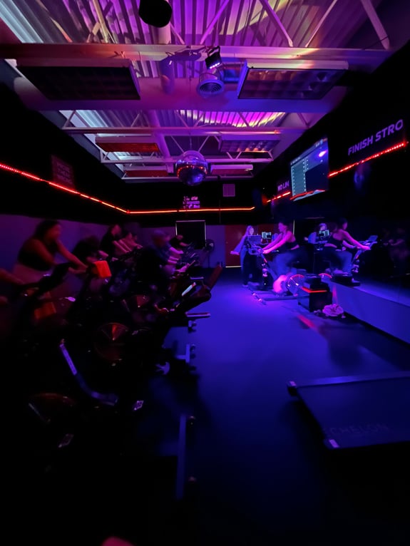 People cycling on stationary bikes in a dimly lit studio with purple lighting.
