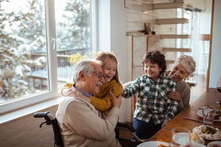 Senior in wheelchair embraced by grandchildren at dining table.