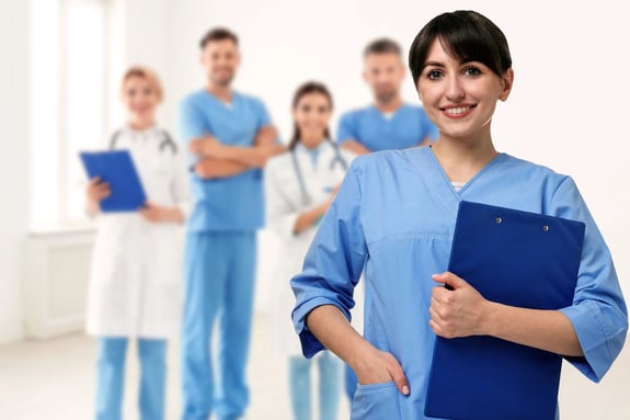 Smiling female nurse in blue scrubs holding a clipboard with team behind.
