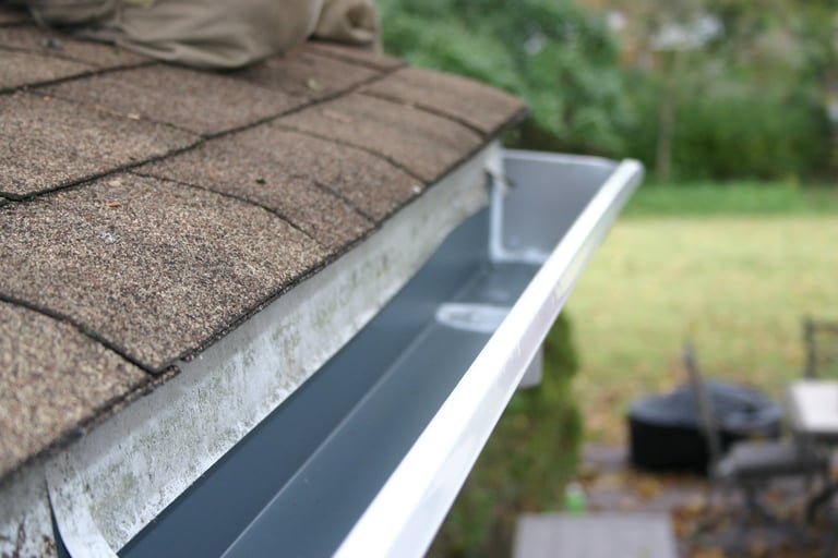 A close-up of a clean, freshly installed house gutter along a roof with brown shingles. The seamless gutter appears free from debris, ready to handle rainwater efficiently.