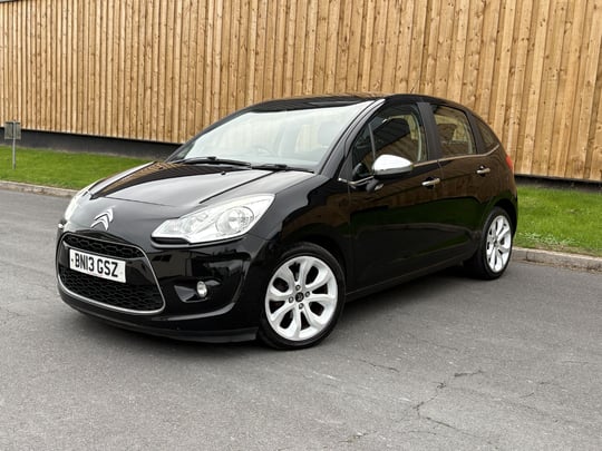 Black Citroën C3 parked on asphalt with wooden fence background.