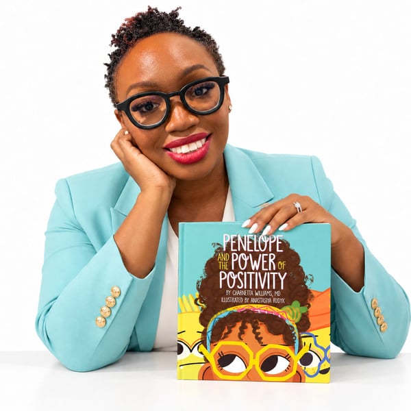 A smiling woman in glasses and blazer holds a children's book.