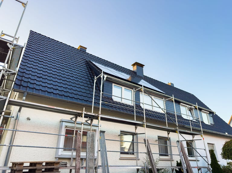 Workers are replacing the roof of a house with scaffolding set up around the building. A clear blue sky is visible in the background, indicating a nice day for the work.
