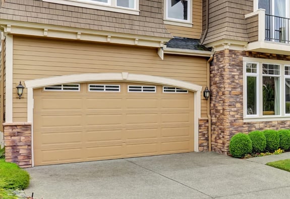 Beige garage door on a two-story house with stone accents and manicured shrubs.