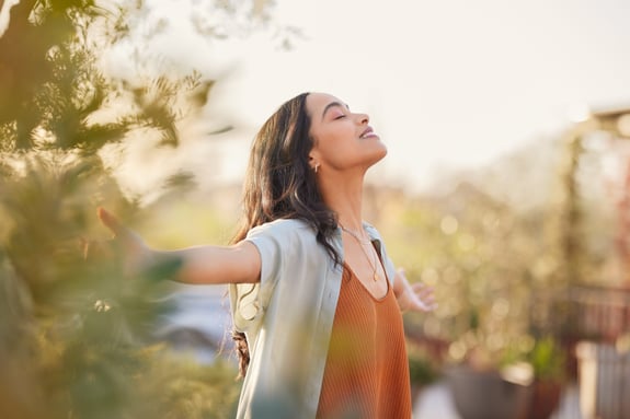 Young latin woman with arms outstretched breathing in fresh air during sunrise at the balcony. Girl enjoying nature while meditating during morning with open arms and closed eyes. Mindful woman relax.