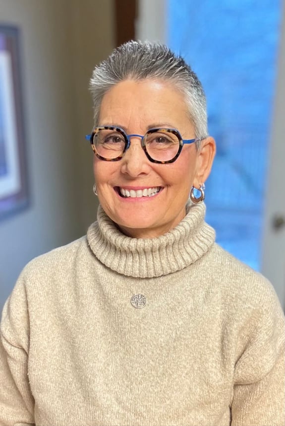 A smiling woman with short gray hair and glasses in a beige turtleneck.