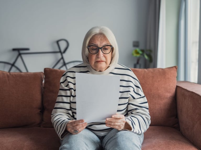 Senior woman with glasses reads a document while seated on a sofa.