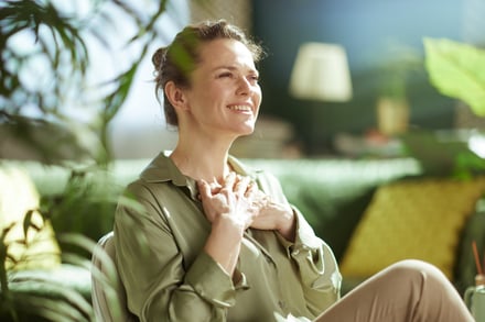 A serene woman in her 40s holds her hands over her heart, expressing gratitude and peace in a beautiful, sunlit room filled with plants.