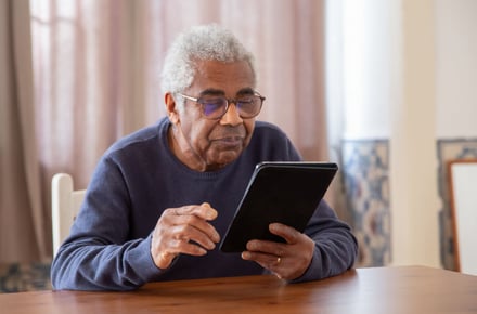 Senior man with glasses using tablet at wooden table indoors.