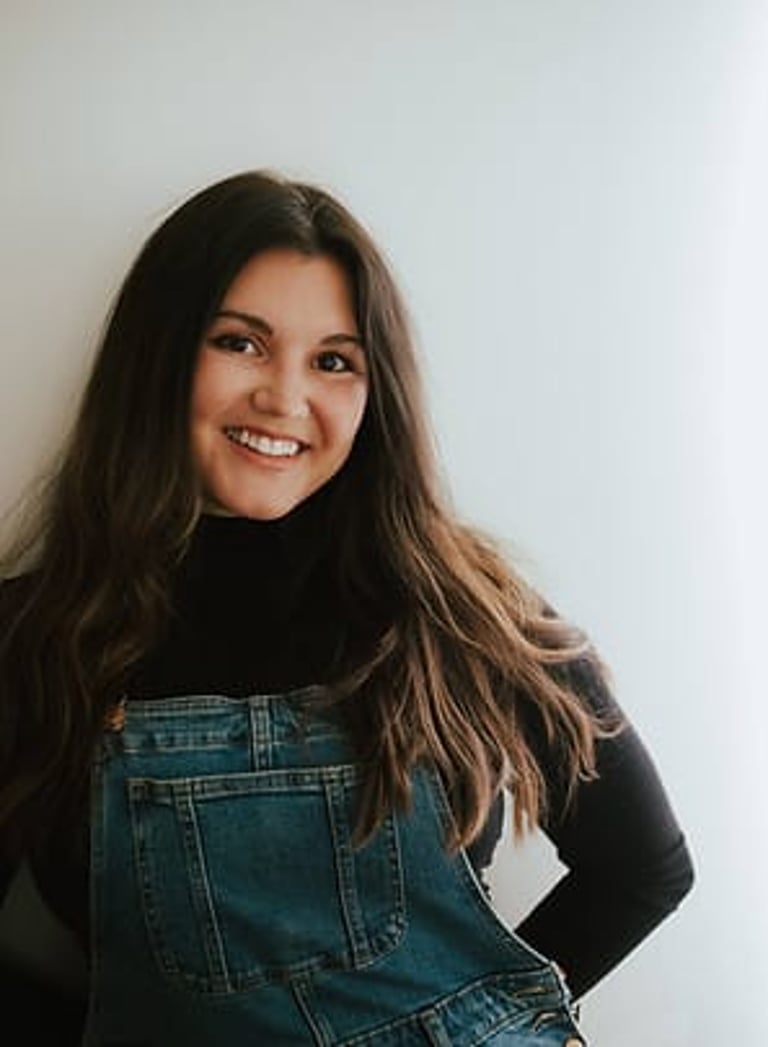 Young woman in denim overalls smiling at camera against plain wall.