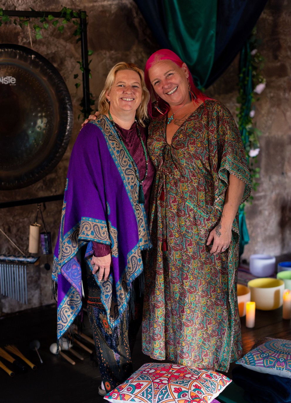 Two smiling women in bohemian attire pose beside a large gong in a decorated studio.