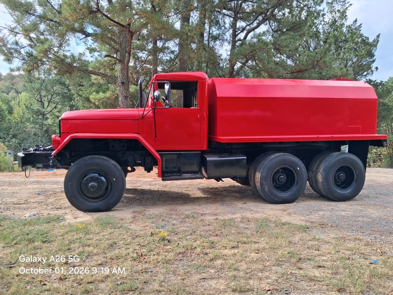 Vintage red tanker truck parked on grass near pine trees.