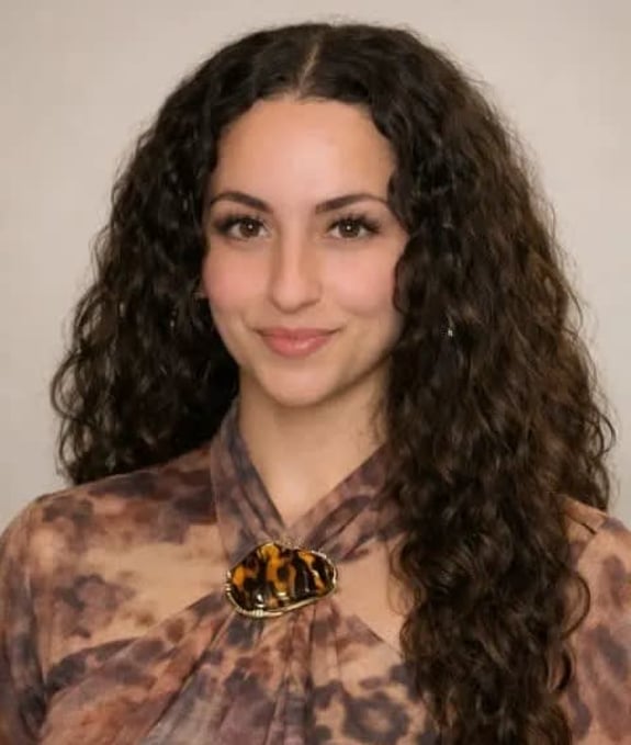 Woman with long curly hair wearing patterned blouse with tortoiseshell brooch.