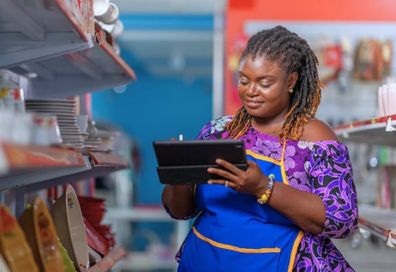 Woman in apron checks inventory on tablet in store aisle.