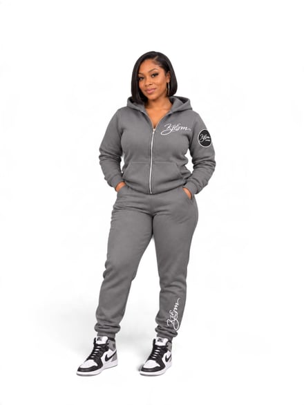 Woman in grey zip-up tracksuit with logo, hands in pockets, standing against white background.