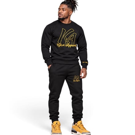 Man in black sweat set with gold logo and yellow sneakers standing against white background.