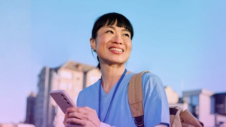 Woman in blue scrubs holding phone outdoors with city buildings behind.